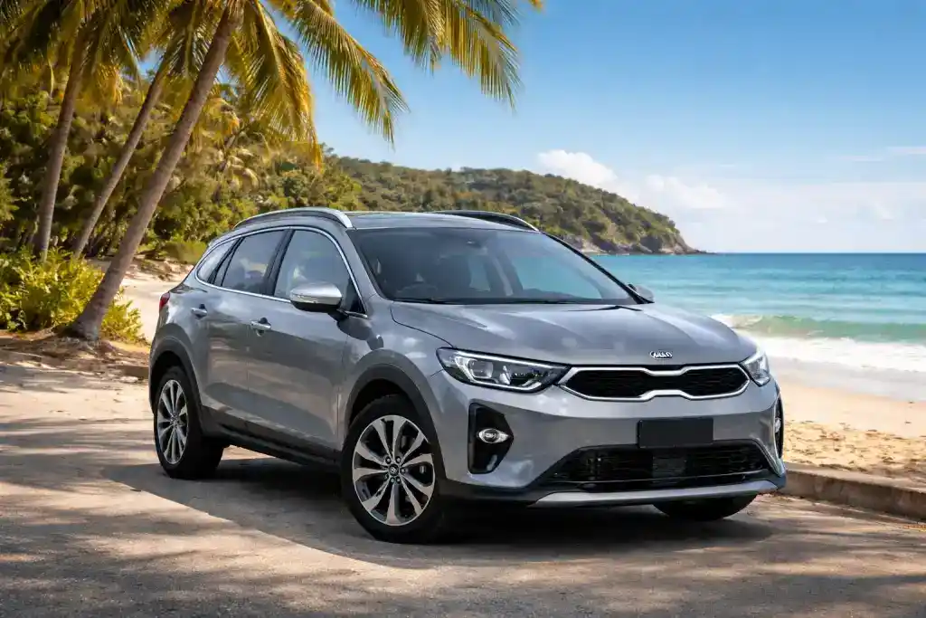 kia-1024x683 1_compressed 1 - Trinitybeachcarrentals.com.au Silver Kia SUV parked on a sunny tropical beach road beside palm trees and blue ocean.