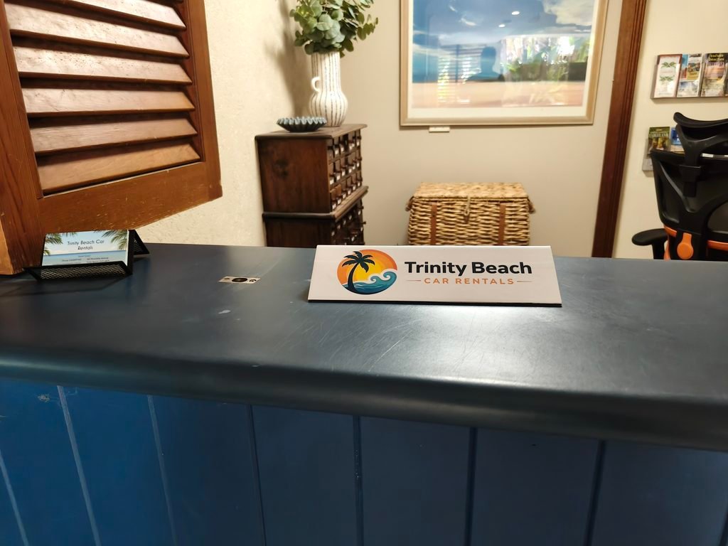 oplus_2097152 - Reception desk with a Trinity Beach Car Rentals sign on a blue counter, wicker basket, wooden cabinet, potted plant, and framed wall art behind it.