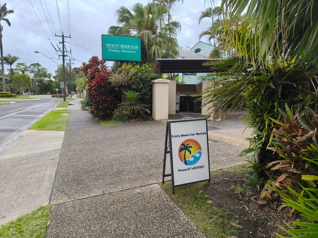 oplus_2097152 - Signboard for Trinity Beach Car Rentals on a tropical street lined with palm plants and bushes.